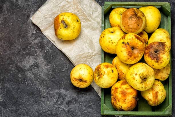 A group of spotted, yellow apples in a green wooden box, with two more apples placed on a piece of brown parchment paper next to it. They are set against a dark, textured background.