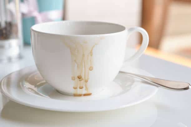 White coffee cup on a saucer with brown stains dripping down the side, a spoon resting beside it.