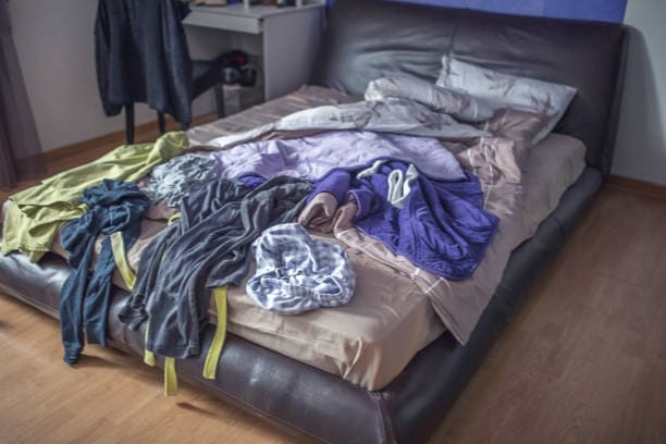 A messy bed with wrinkled sheets and various clothes scattered on top in a room featuring a small desk and chair in the background.
