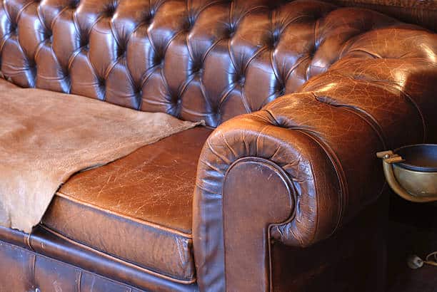 Close-up of a vintage brown leather Chesterfield sofa with tufted cushions and a rolled armrest. A suede throw lies across the seat.