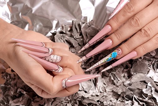 Hands with long, pointed acrylic nails in pink and floral designs. They are shown against a textured gray background.