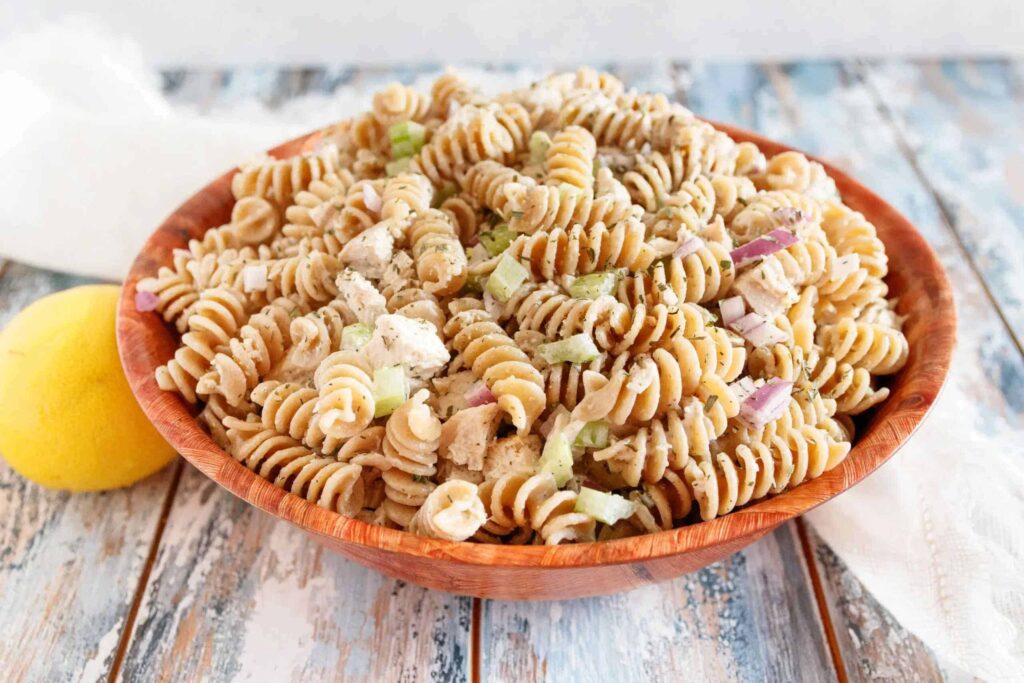 A wooden bowl of rotini pasta salad with diced chicken, celery, and red onion. A lemon is placed beside the bowl on a rustic wooden surface.