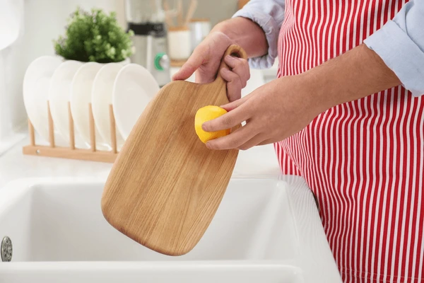 Person cleaning a wooden cutting board with half a lemon over a kitchen sink.