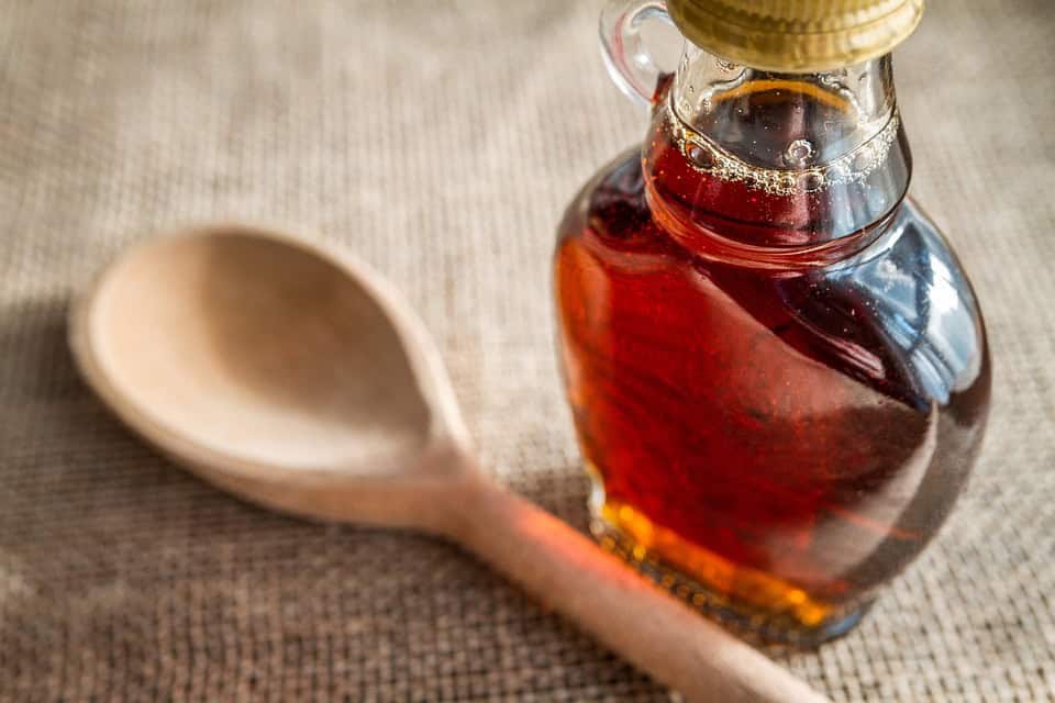 A glass bottle of syrup with a golden cap sits next to a wooden spoon on a burlap surface.