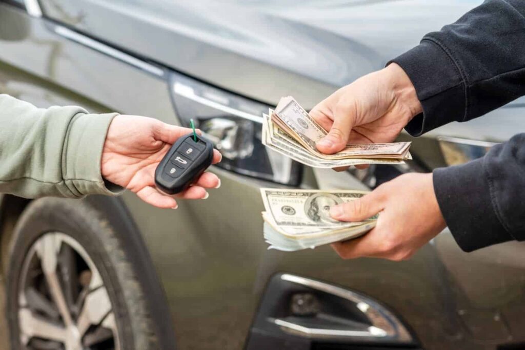 Close-up of two people exchanging a car key and cash in front of a silver vehicle.