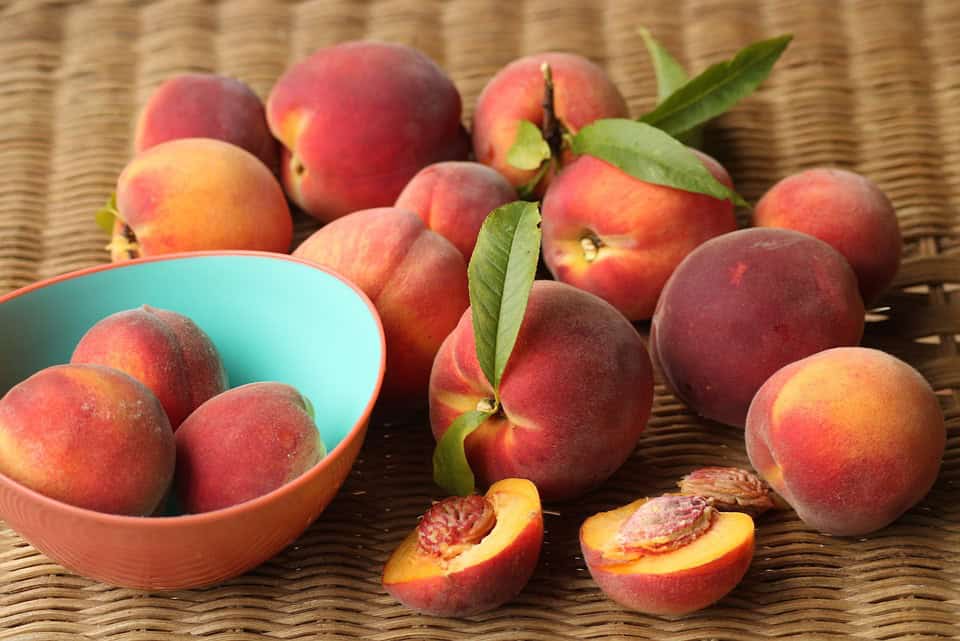 Peaches scattered on a woven surface, some in a bowl. One peach is cut open, showing the pit. Green leaves are attached to a few peaches.