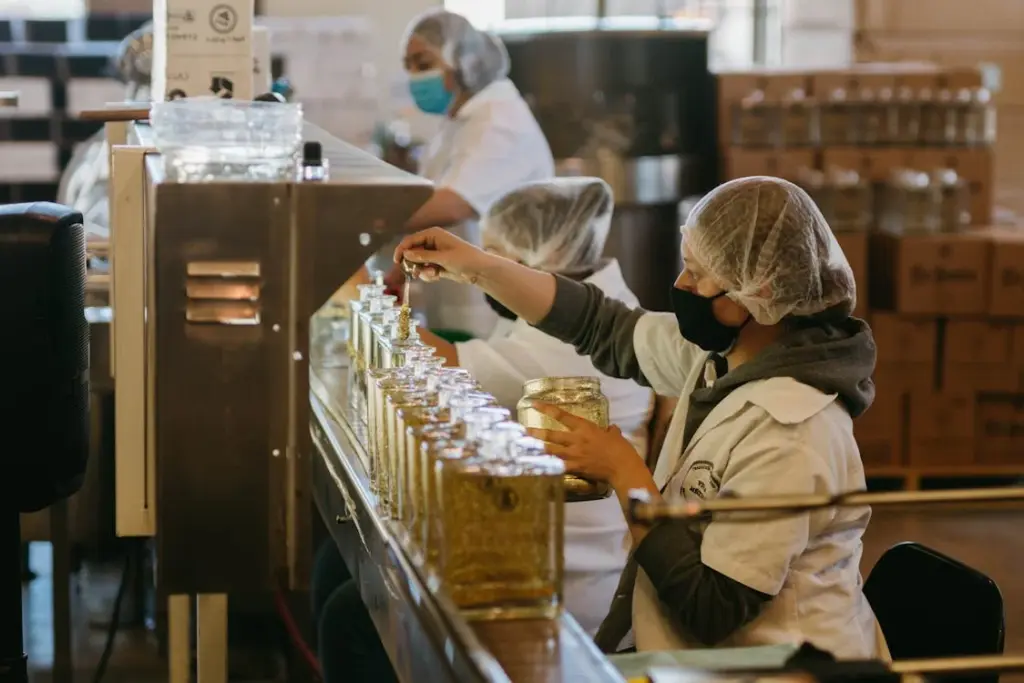 Workers in a factory, wearing hairnets and masks, are filling glass bottles with liquid at a production line.