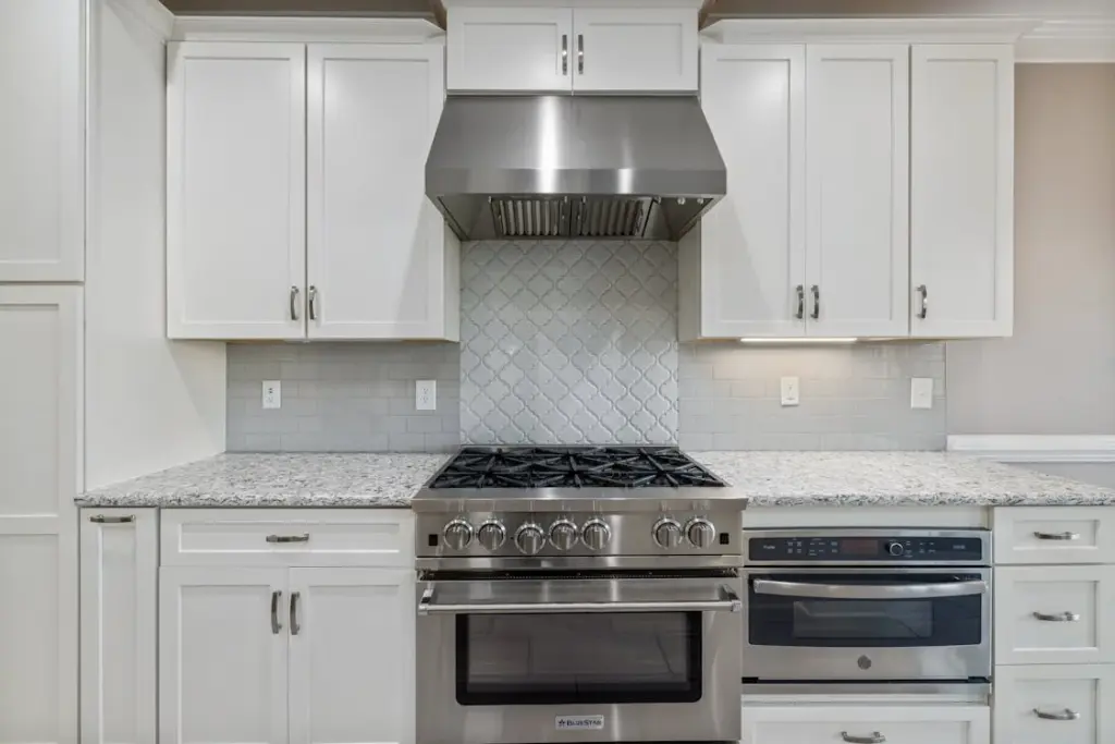 A modern kitchen with white cabinets, a stainless steel range hood, a gas stove, and a tiled backsplash above a granite countertop.