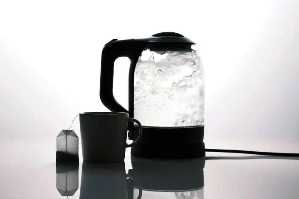 A glass kettle with boiling water is next to a mug and a tea bag on a reflective surface.