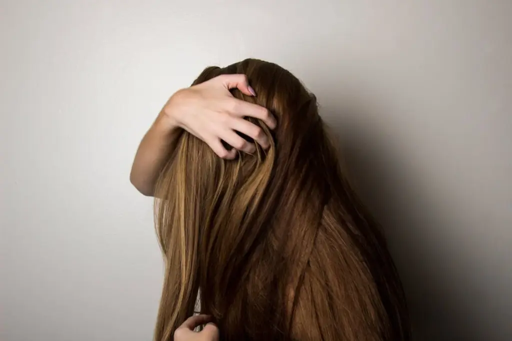 A person with long brown hair faces a blank wall, partially covering their face with their hair. One hand is holding the top of their head.
