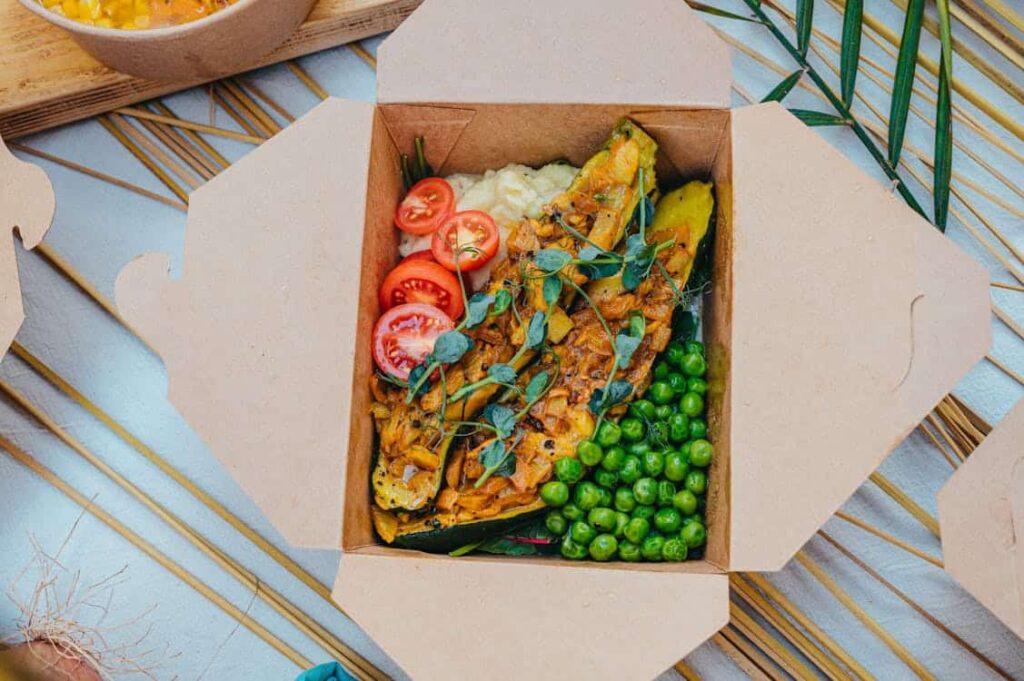 A cardboard box contains a meal with grilled vegetables, peas, sliced cherry tomatoes, and a creamy sauce, garnished with herbs.