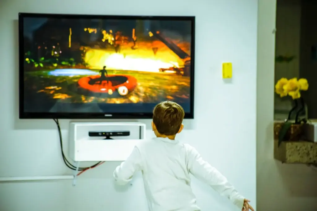 A child in a white shirt watches a video game on a wall-mounted TV, standing closely and engaging with the screen.