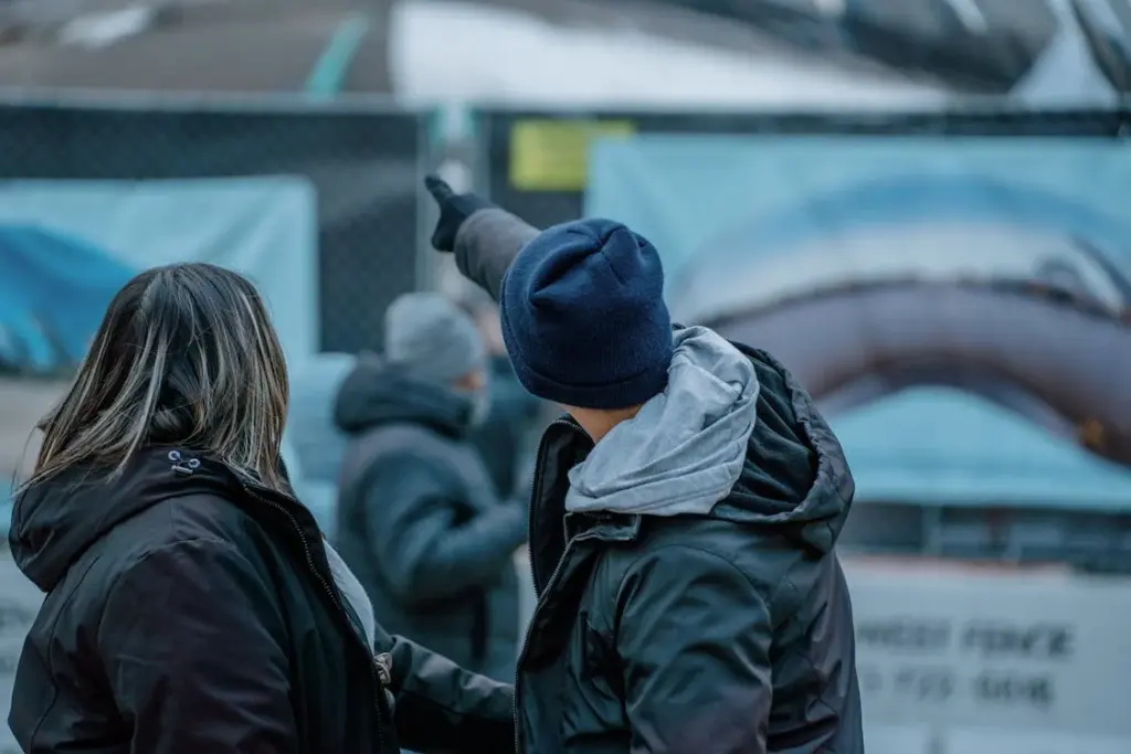 Two individuals wearing winter clothing stand near a construction site. One person points towards the site in the background.