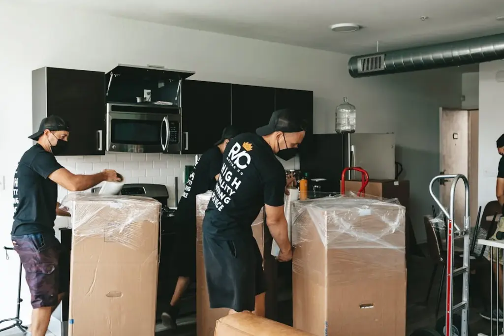 Three movers in black shirts and caps, wrapping large appliances in plastic, work inside a modern kitchen with stainless steel appliances and dark cabinets.