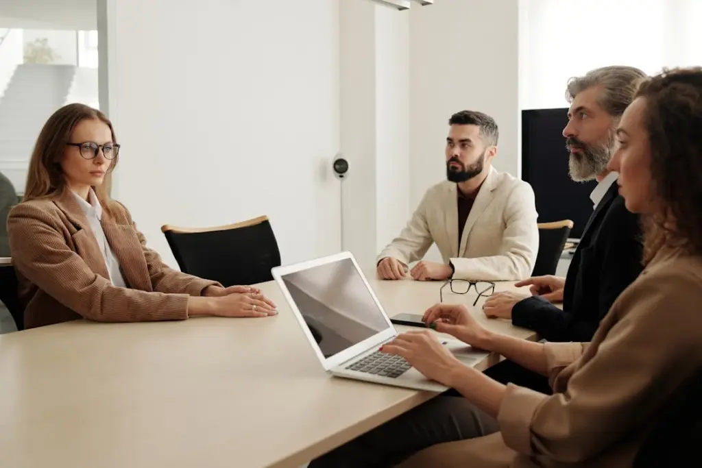 A woman sits across from three people at a conference table. One person holds a laptop. They appear to be engaged in a meeting.