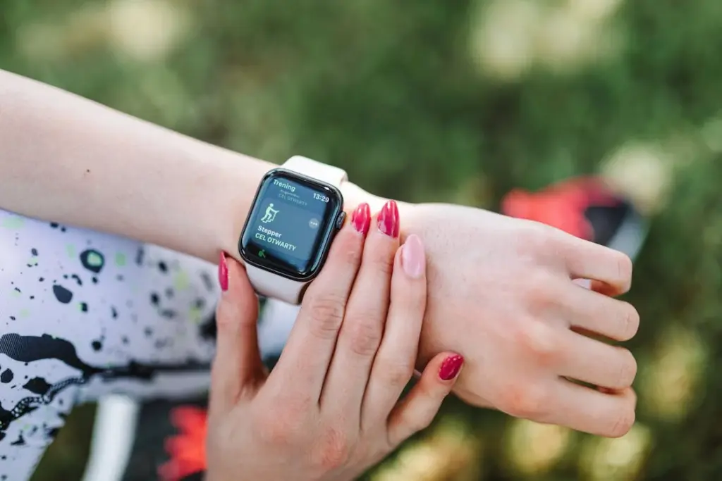 A person wearing a smartwatch on their left wrist, displaying a step count. They have red and pink nails, and a blurred grassy background is visible.