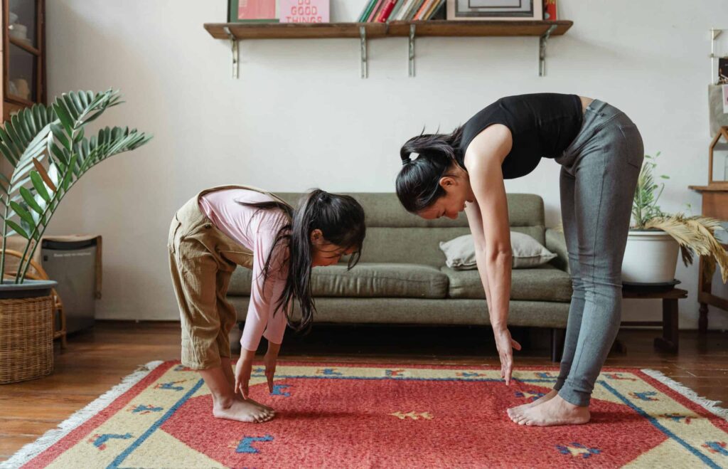 A woman and child practice yoga in a living room, bending forward with straight legs and arms reaching toward the floor on a red patterned rug.