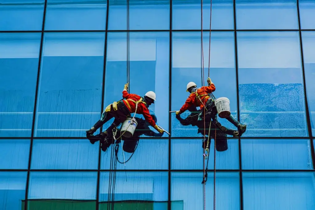 Two workers in safety gear clean a large glass building using ropes and tools.