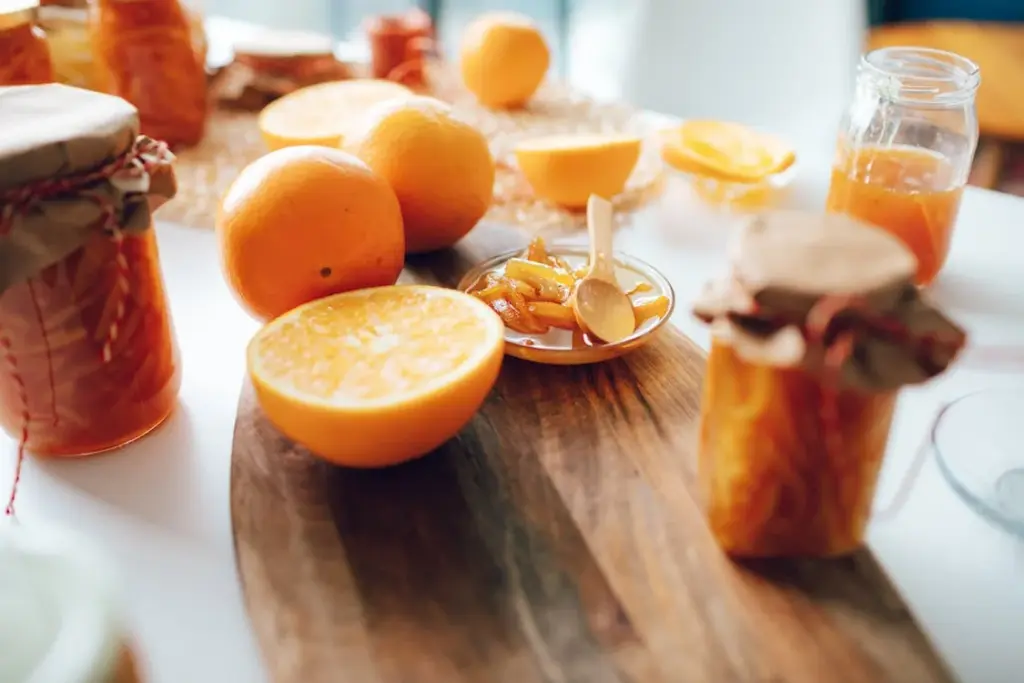 A wooden board with oranges, orange marmalade in jars, and a small dish with marmalade, on a white table.