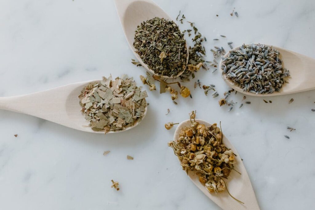 Four wooden spoons on a marble surface filled with different dried herbs and flowers, including lavender, chamomile, and other leafy herbs.