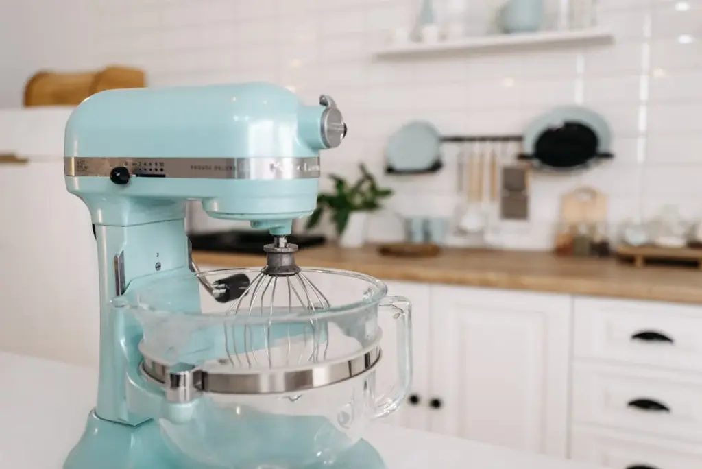 A light blue stand mixer with a whisk attachment and glass bowl on a kitchen counter. White tile backsplash and wooden shelves are in the background.