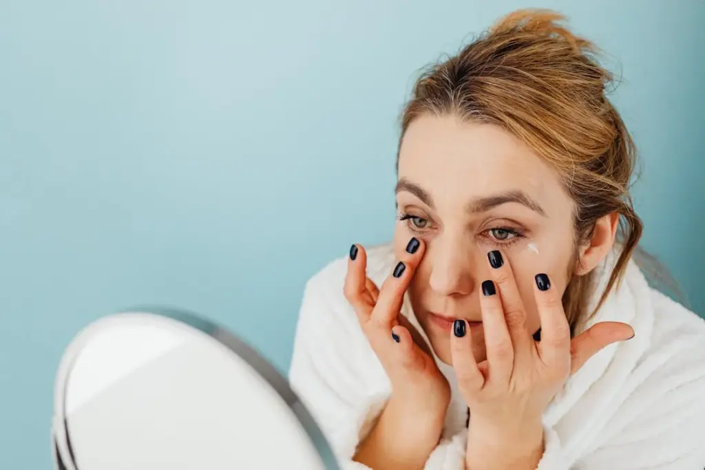 A woman applies eye cream with her fingers while looking in a mirror, against a light blue background.