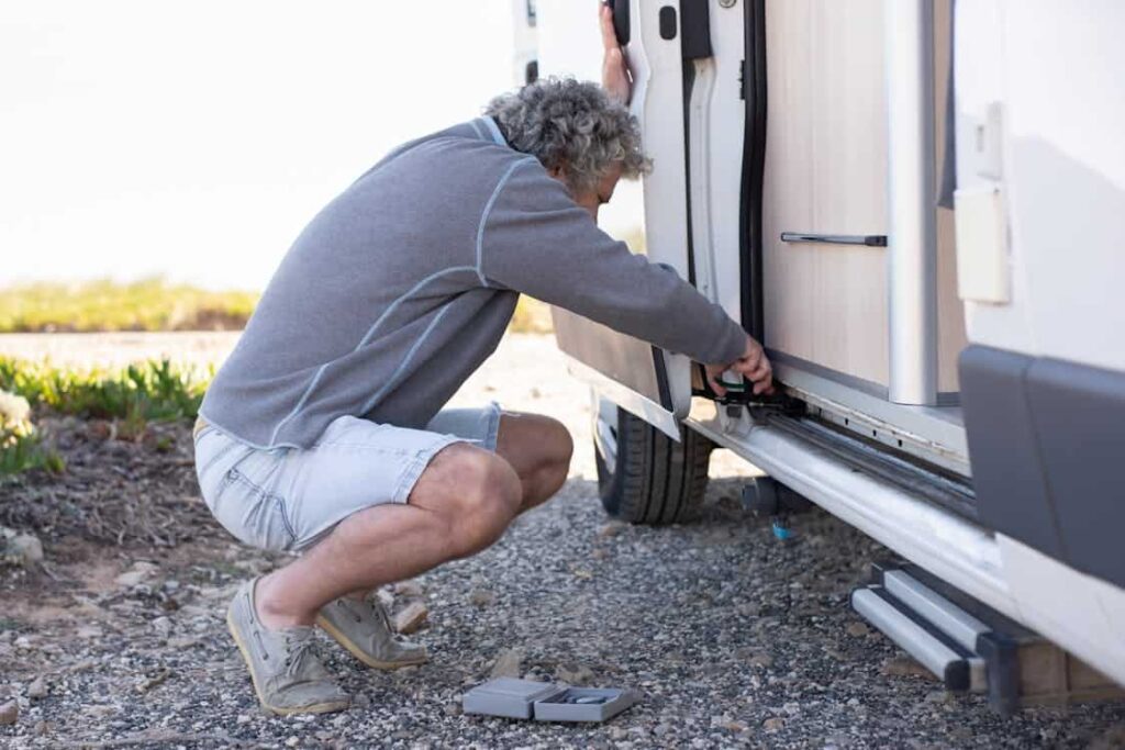 Man in shorts and a gray sweater kneeling on rocky ground, fixing or inspecting an open compartment on the side of a vehicle.