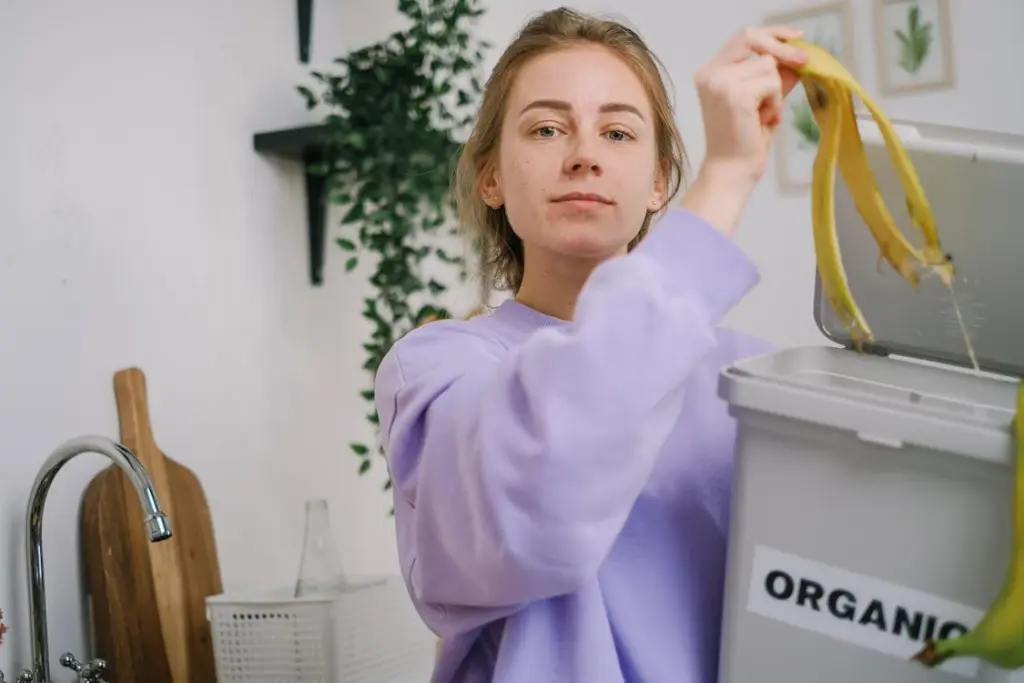 Person in a purple sweater disposing of banana peels into an organic waste bin in a kitchen.