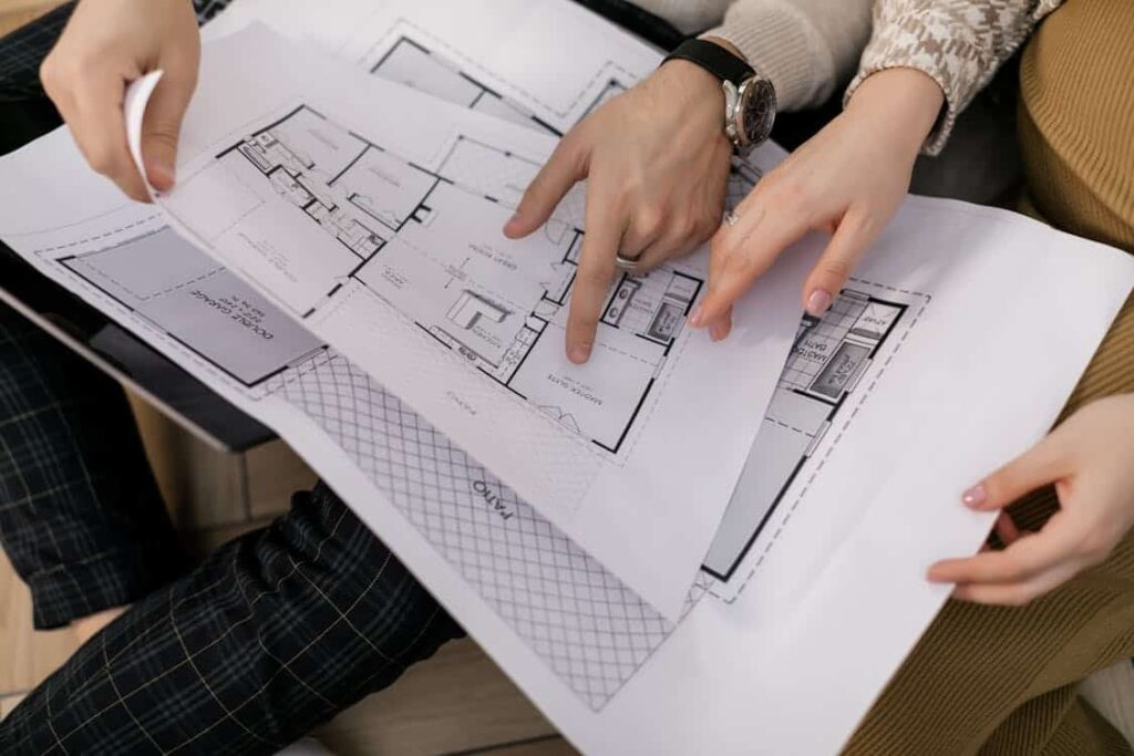 Two people examine architectural floor plans spread on a table, with one person pointing at specific details.