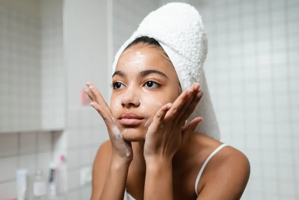 Person applying skincare cream on face in a tiled bathroom, with a towel wrapped around their head.