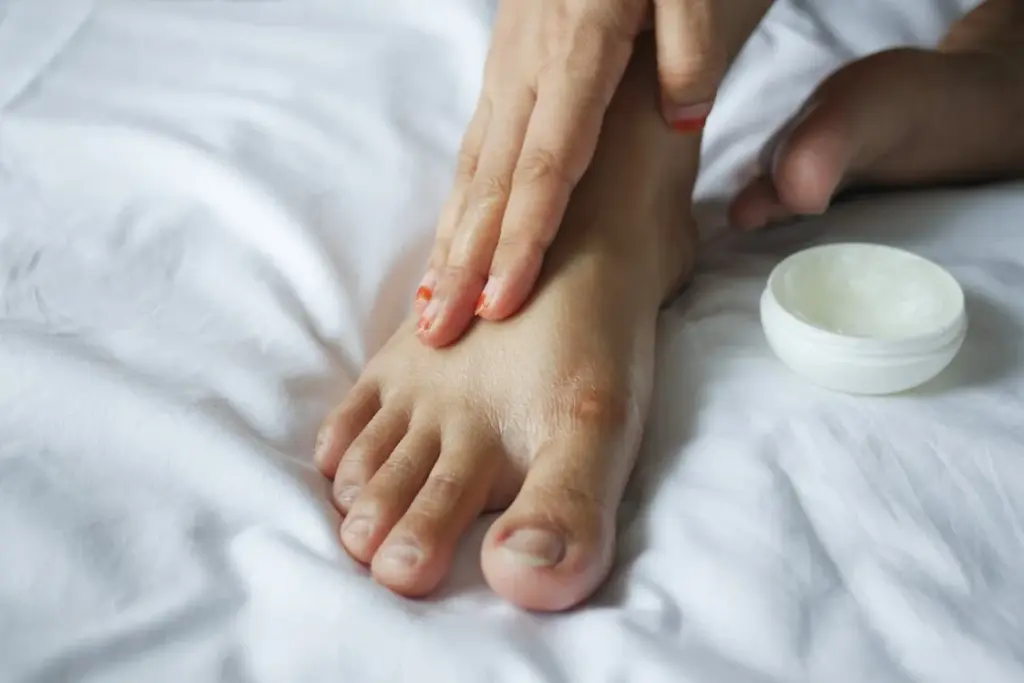 A person applies cream to their foot on a white bed. A small jar of cream is nearby.