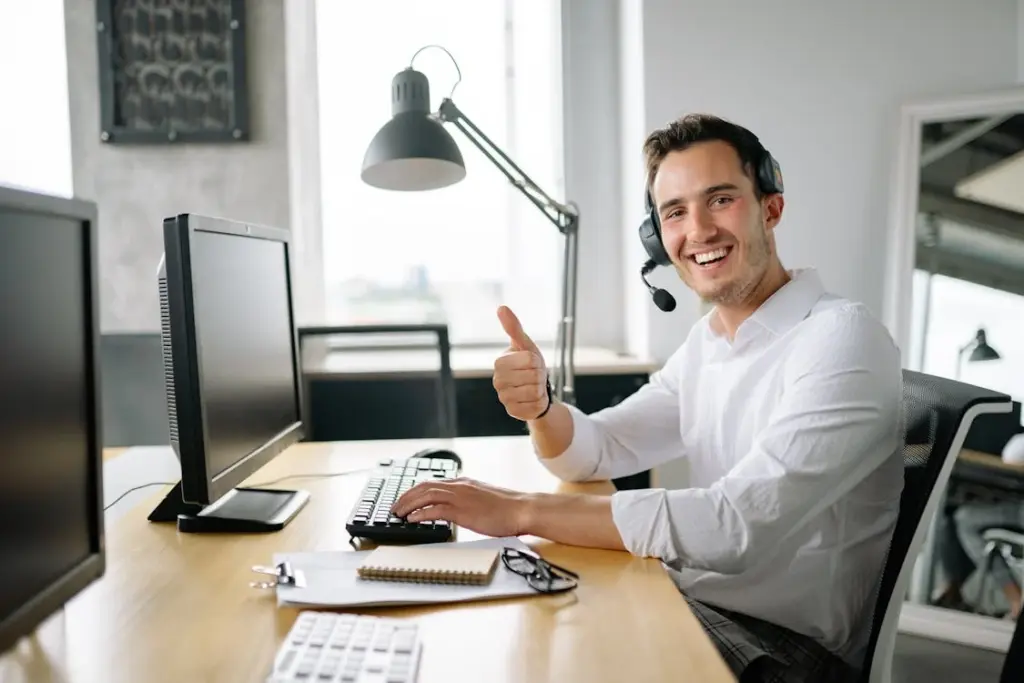 Person at a desk with dual monitors, wearing a headset, smiling, and giving a thumbs-up.