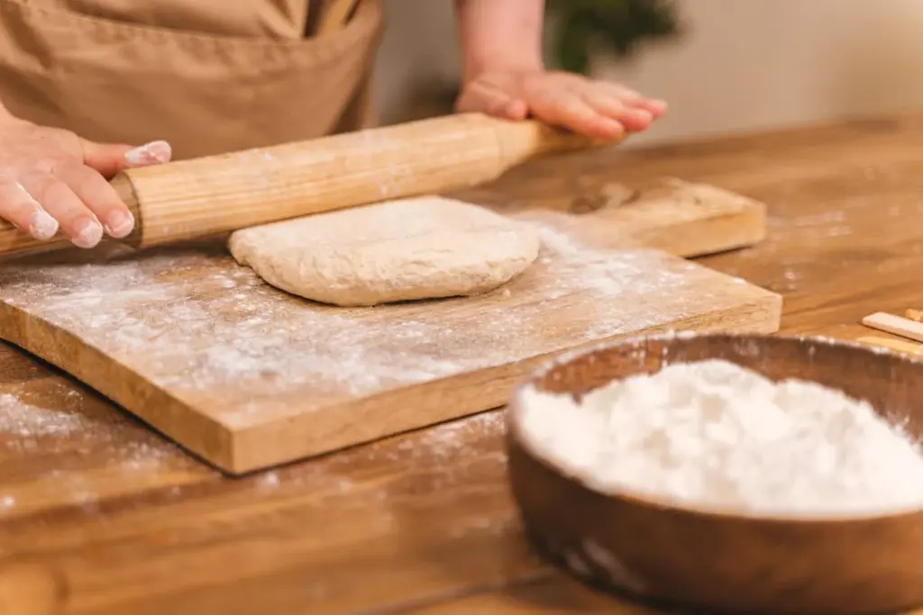 Person wearing an apron uses a rolling pin on dough placed on a floured wooden board. A bowl of flour is in the foreground.