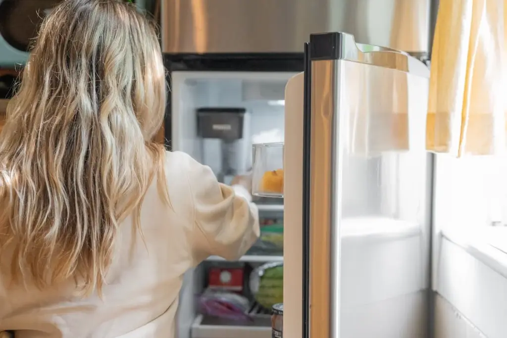 A person with long hair is looking inside an open refrigerator, standing in a brightly lit kitchen.