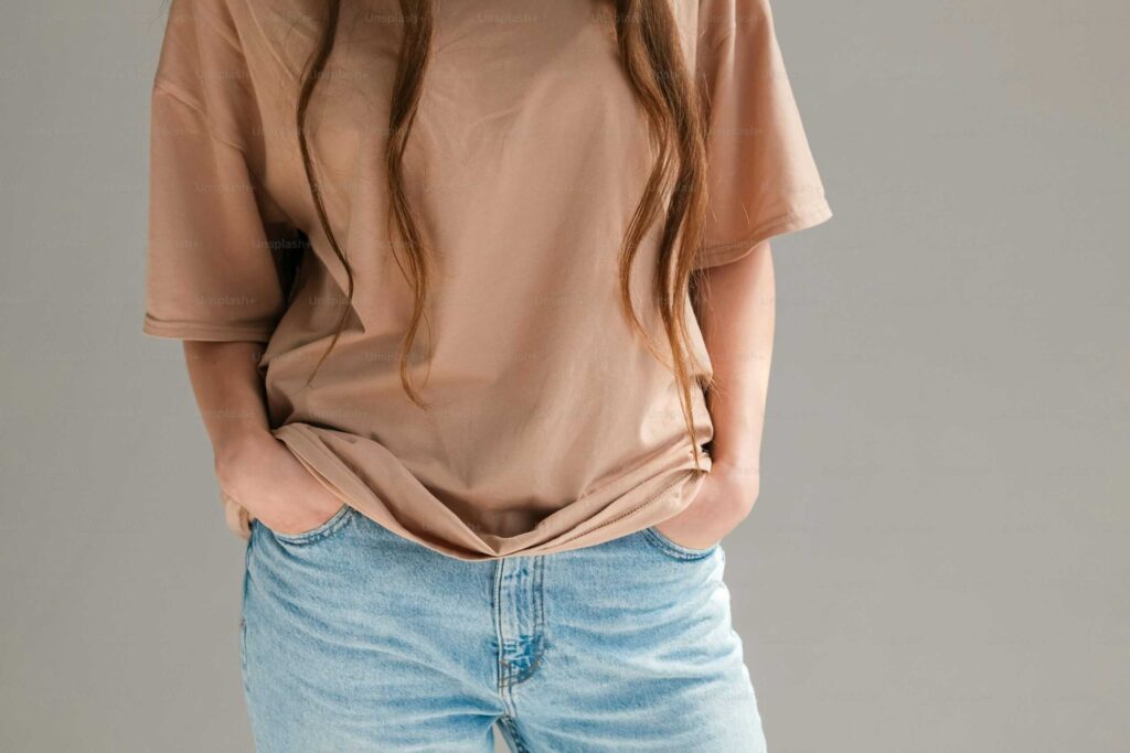 A person wearing a beige t-shirt and light blue jeans stands with hands in pockets against a neutral background.