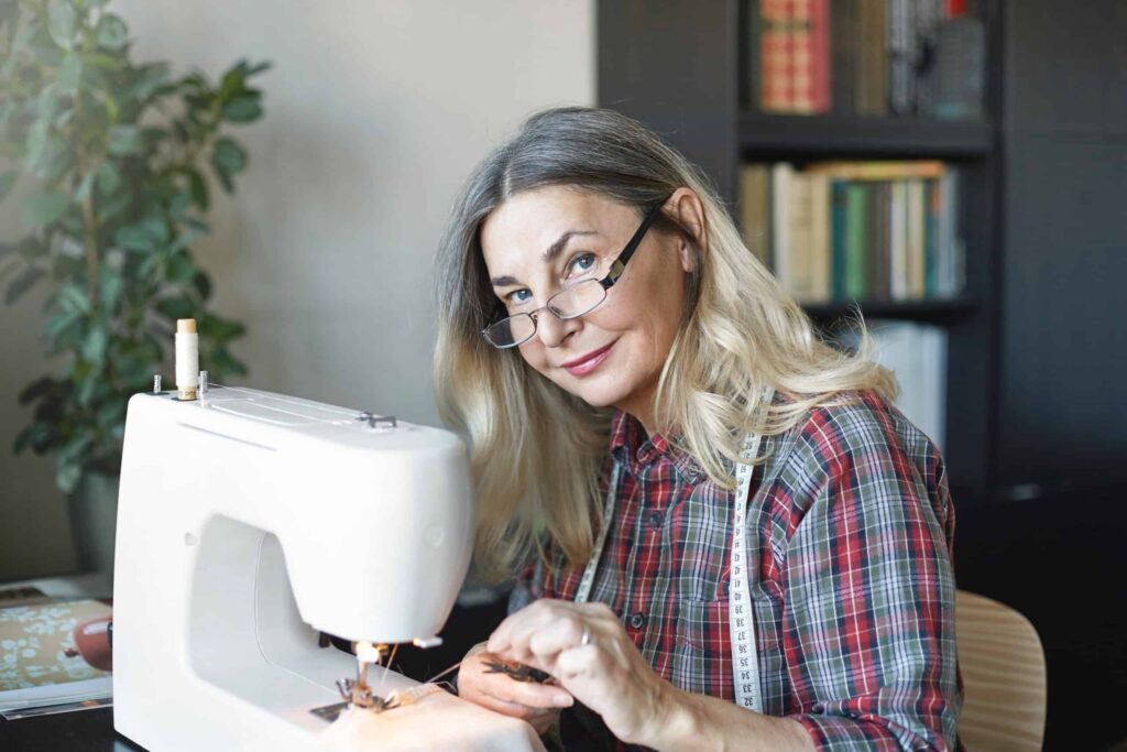 A woman with glasses and a plaid shirt uses a sewing machine, surrounded by books and a potted plant.