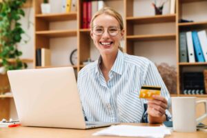 A person wearing glasses and a striped shirt smiles while holding a credit card and sitting at a desk with a laptop, coffee mug, and paperwork. Shelves with books and plants are in the background.