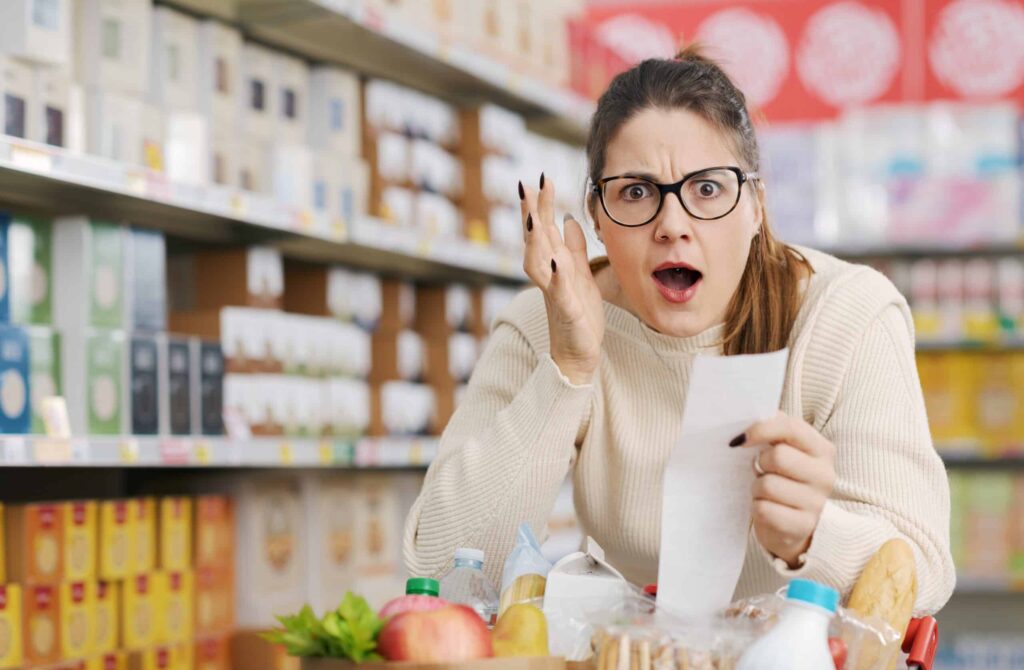 A shocked woman holds a shopping receipt in a grocery store aisle.