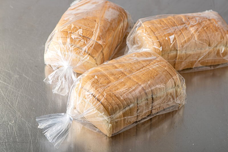 Three loaves of sliced white bread in plastic bags on a metal surface.
