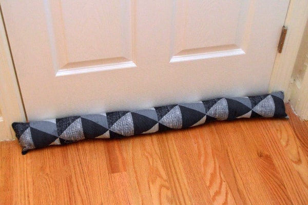 A draft stopper with a geometric pattern is placed at the base of a closed white door on a wooden floor.