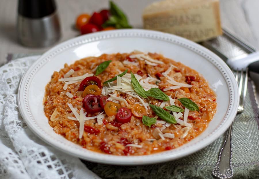 A plate of tomato risotto garnished with cherry tomatoes, basil leaves, and shredded cheese, placed on a tablecloth with a napkin, salt shaker, and a wedge of cheese in the background.