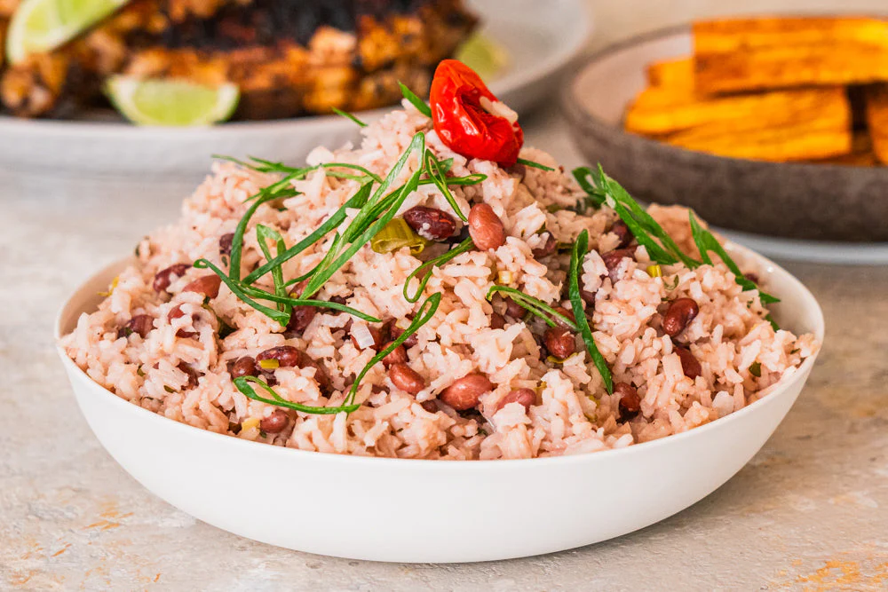 A bowl of rice and beans garnished with green herbs and a red pepper. Plates of grilled food and plantains are in the background.