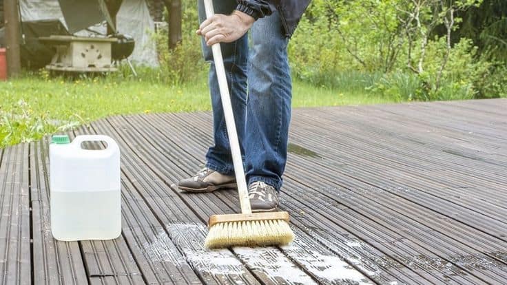 Person scrubbing a wooden deck with a brush. A container of cleaning solution is nearby. Garden and outdoor furniture are visible in the background.