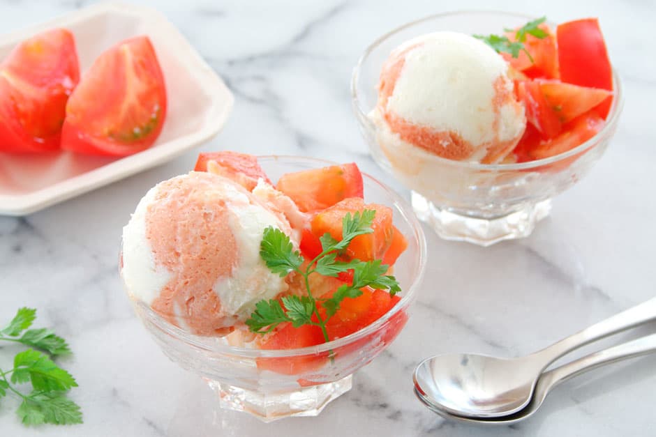 Two glass bowls with tomato sorbet and diced tomatoes, garnished with parsley, on a marble surface. A plate with sliced tomatoes and a spoon are nearby.
