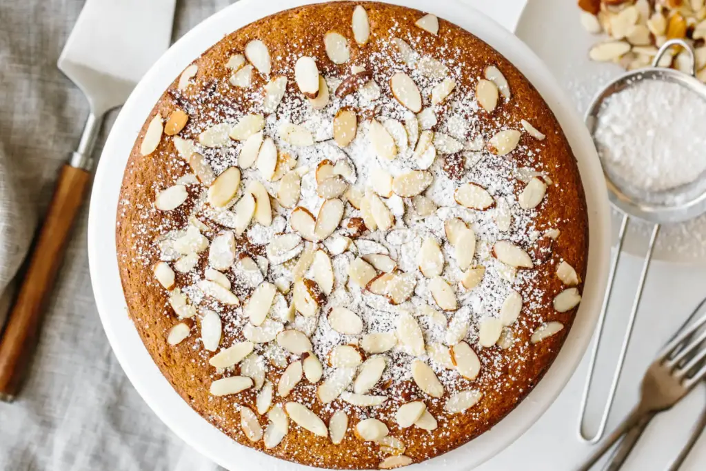 A round cake topped with powdered sugar and almond slices on a white plate. Baking tools and a small bowl of almonds are nearby.