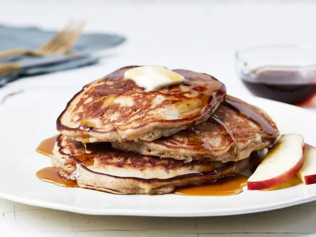 A stack of pancakes topped with a pat of butter and syrup on a white plate, accompanied by apple slices and a bowl of syrup.