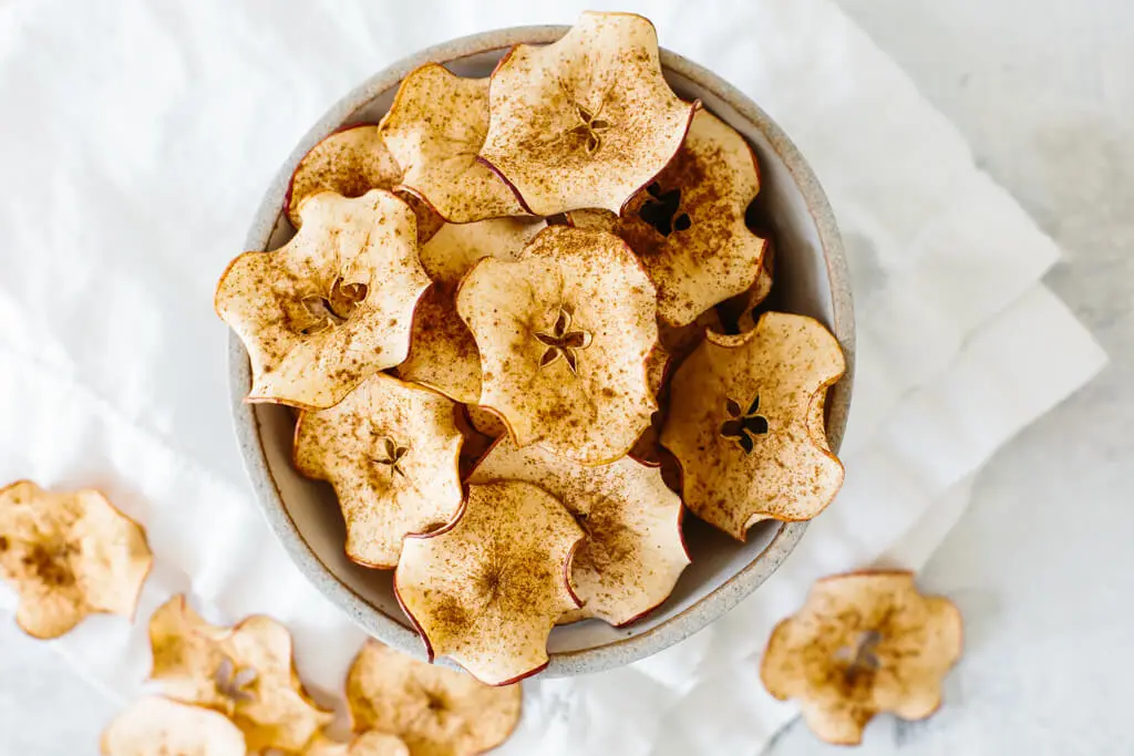 A bowl of cinnamon-dusted apple chips is placed on a white napkin, with a few chips scattered around.