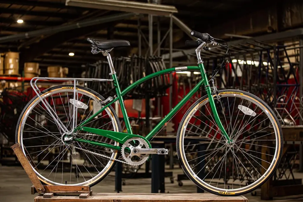A green bicycle with a metal frame and black seat is displayed indoors on a wooden stand. Industrial background with racks and equipment.