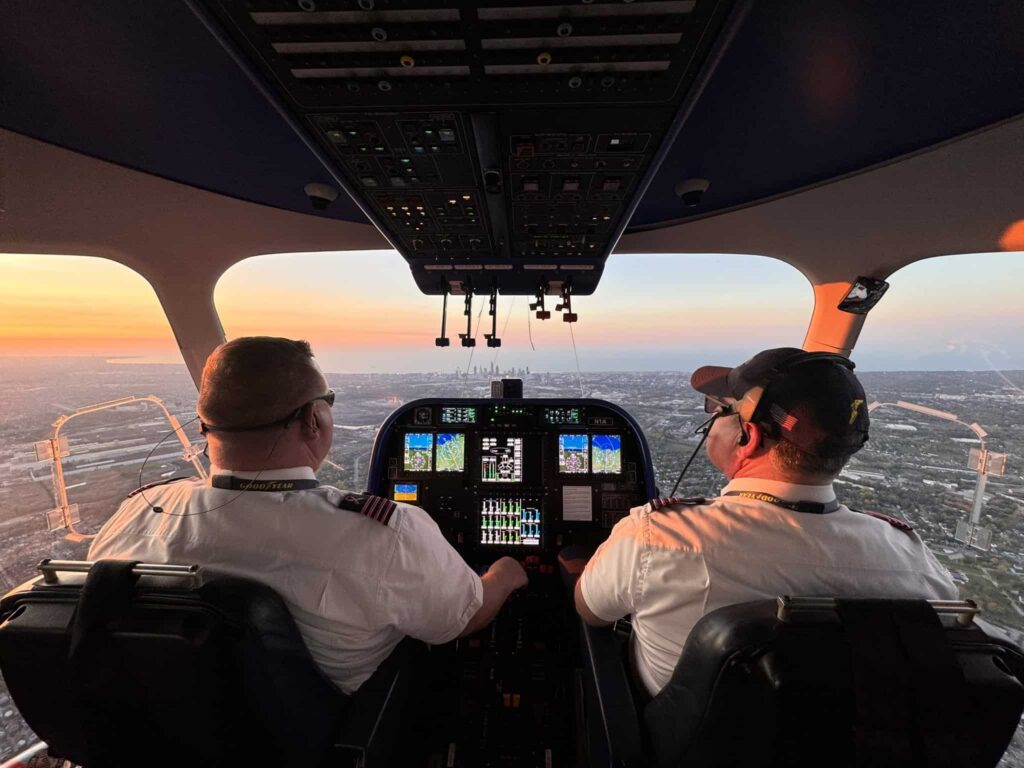 View from behind two pilots in a cockpit, flying over a city with a sunrise or sunset visible through the windows.