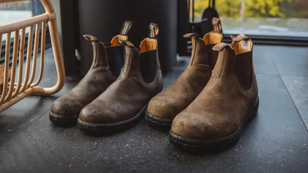 Two pairs of brown leather boots with black soles are placed side by side on a dark tiled floor near a wooden chair.